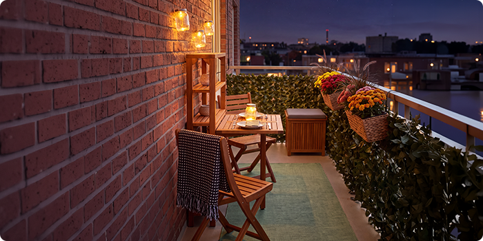 Malý balkon s dřevěnými židlemi a stolem, osvětlený lucernami a svíčkami, zdobený květinami v květináčích a zelení, s nočním výhledem na městské panorama.